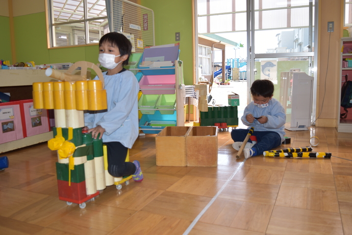 ブロックで作った車に乗る幼児と、筒に黒と黄色のビニールテープを巻いている幼児