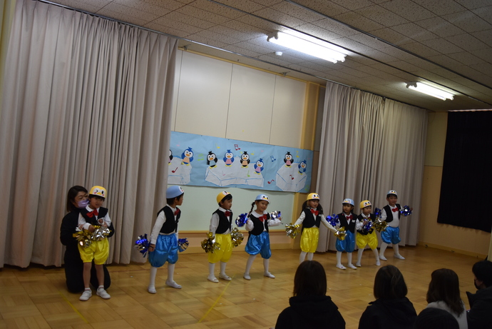 青や黄色のズボンと黒のタキシードでポンポンを持って踊る園児たち